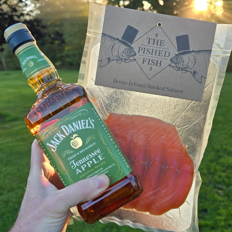 A hand holds a bottle of Jack Daniel's Tennessee Apple bourbon whiskey and a clear plastic bag of smoked salmon slices in front of an outdoor grassy background with sunlight filtering through trees. The bag label reads 'THE FISHED FISH' and 'Booze Infused Smoked Salmon'.